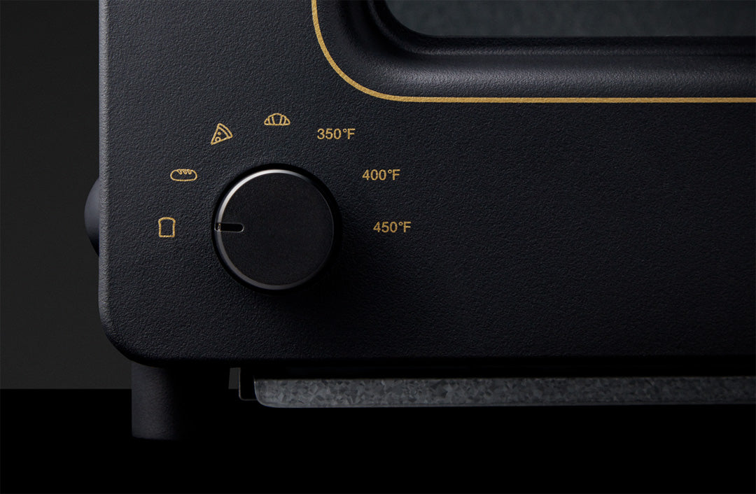 Close-up of a black toaster oven control panel with gold markings. The temperature dial is surrounded by icons for different food types, including toast, bread, croissants, and pizza. Temperature settings range from 350°F to 450°F. The toaster oven has a sleek, minimalist design with a matte black finish and subtle gold detailing.