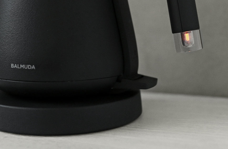 Close-up of the base and handle of a matte black electric kettle by BALMUDA, with the brand name visible and a soft orange light glowing inside the transparent spout tip.
