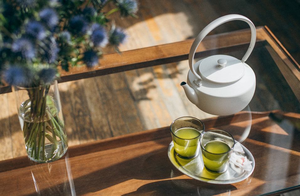 A minimalist white MoonKettle and two glasses of vibrant green tea rest on a glass coffee table, bathed in warm sunlight beside a vase of blue flowers.