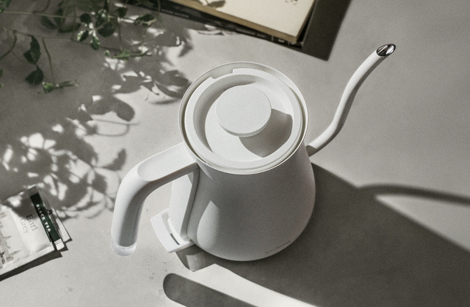 Overhead view of a minimalist white electric kettle with a gooseneck spout placed on a sunlit concrete surface, surrounded by soft shadows and a few tea-related items.
