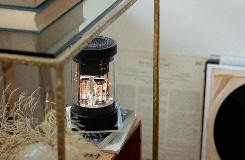 BALMUDA The Speaker placed on a stack of books inside a glass-shelved cabinet with vintage papers in the background.