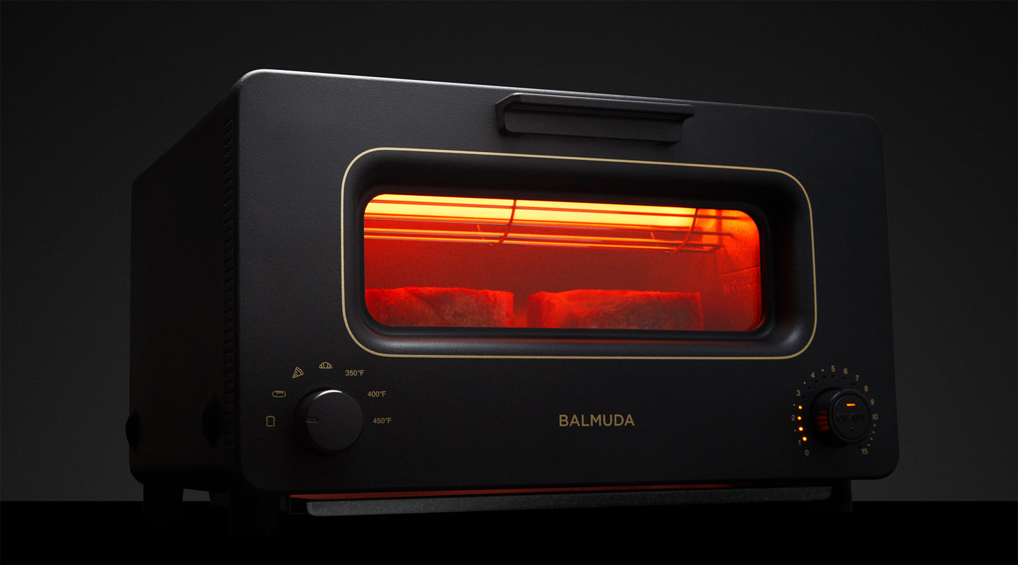 A sleek black BALMUDA The Toaster with a gold-trimmed front panel, glowing red from the heating elements inside. Two slices of bread are toasting, visible through the window. The Toaster features a minimalist design with temperature and timer dials on the front.