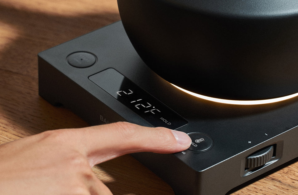 A close-up of a hand pressing the temperature hold button on the BALMUDA MoonKettle, with its digital display reading 212°F and a soft light glowing beneath the kettle.