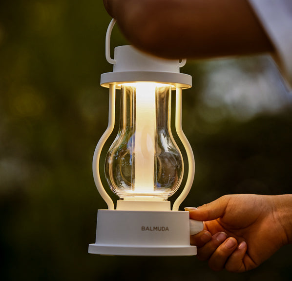 A person’s hand adjusting the brightness of a white BALMUDA lantern glowing warmly outdoors in soft evening light.