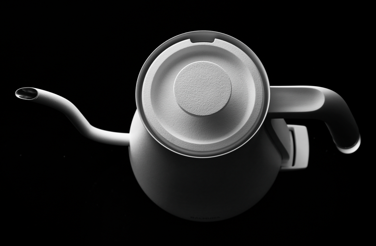 A white BALMUDA kettle viewed from the top against a black background, highlighting its round lid and curved spout.