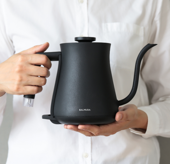 A person in a white shirt holds a black BALMUDA electric kettle with both hands, showing its minimalist design.