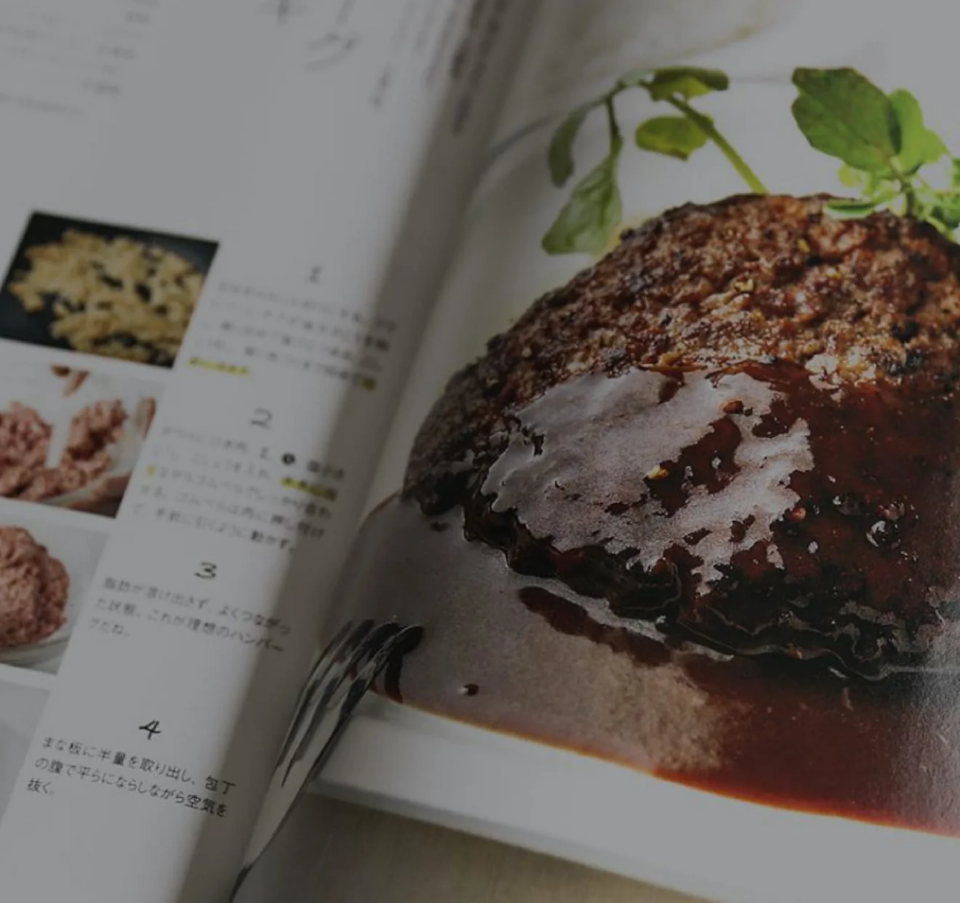 A close-up view of an open cookbook featuring a photo of a hamburger steak covered in sauce, with text and step-by-step images on the opposite page.