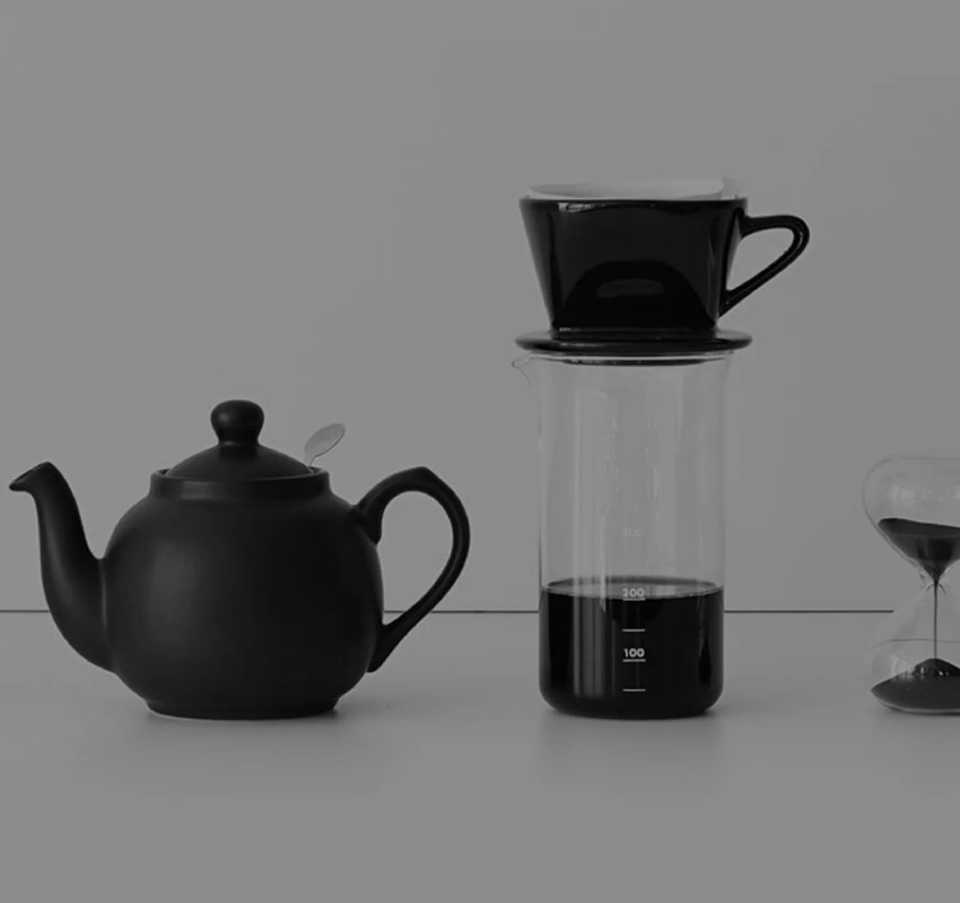A black teapot, glass coffee server with a black dripper on top, and an hourglass timer arranged on a white surface against a neutral background.