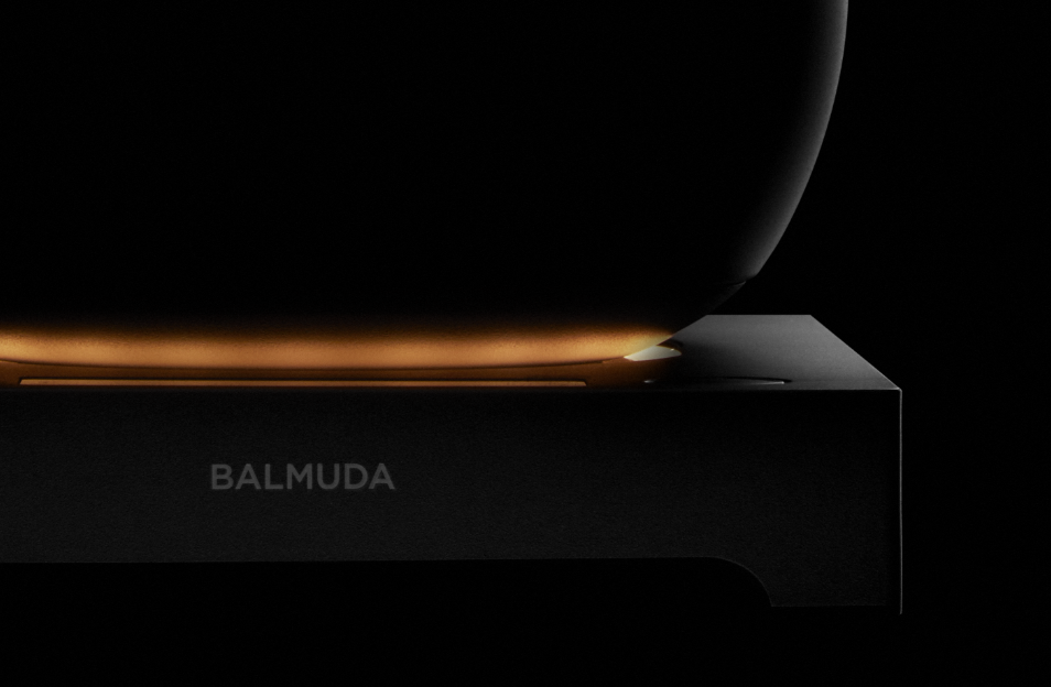 A dramatic close-up of the BALMUDA MoonKettle’s induction base, featuring a soft golden glow beneath the black kettle against a dark background.