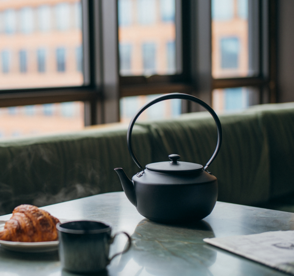 A black MoonKettle sits on a marble table beside a steaming cup and a flaky croissant, with soft natural light filtering through large windows in the background.
