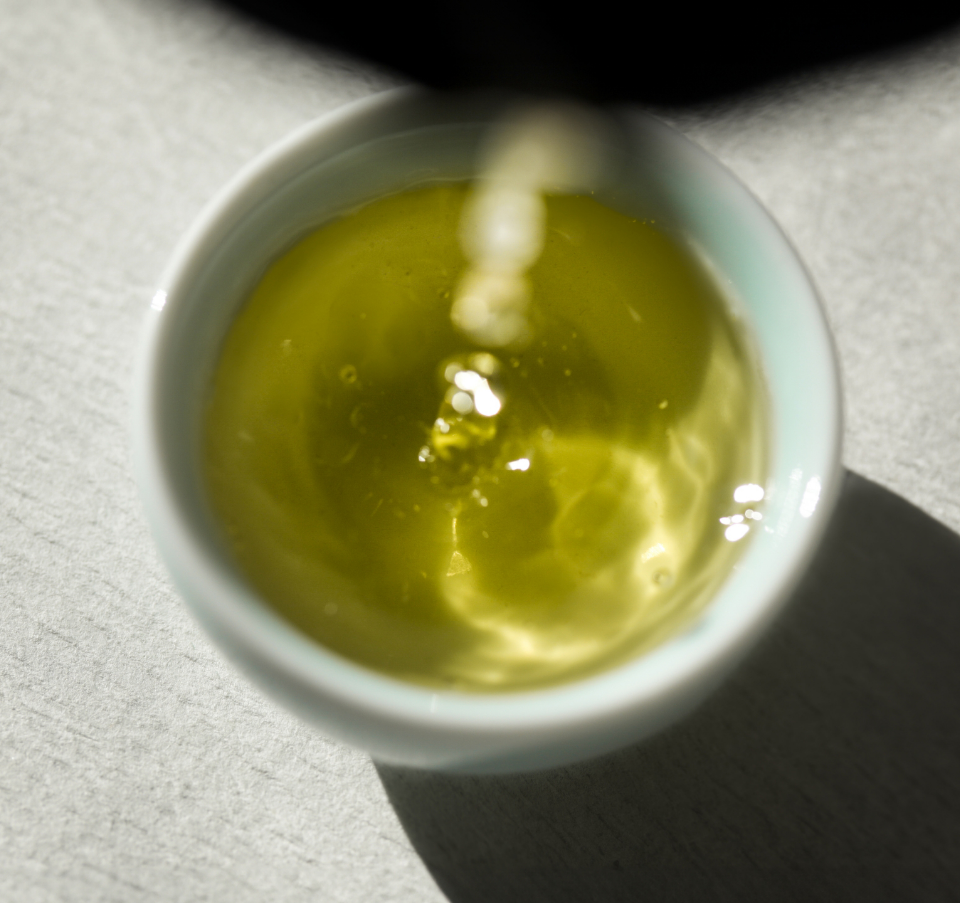 A close-up of vibrant green tea being poured into a white ceramic cup, capturing the ripples and light reflections in the beautiful tea.