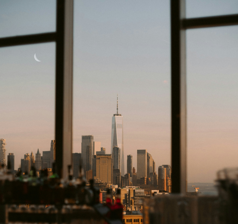 A stunning view of the New York City skyline, framed by a window, with One World Trade Center standing tall against a pastel sky and a crescent moon.