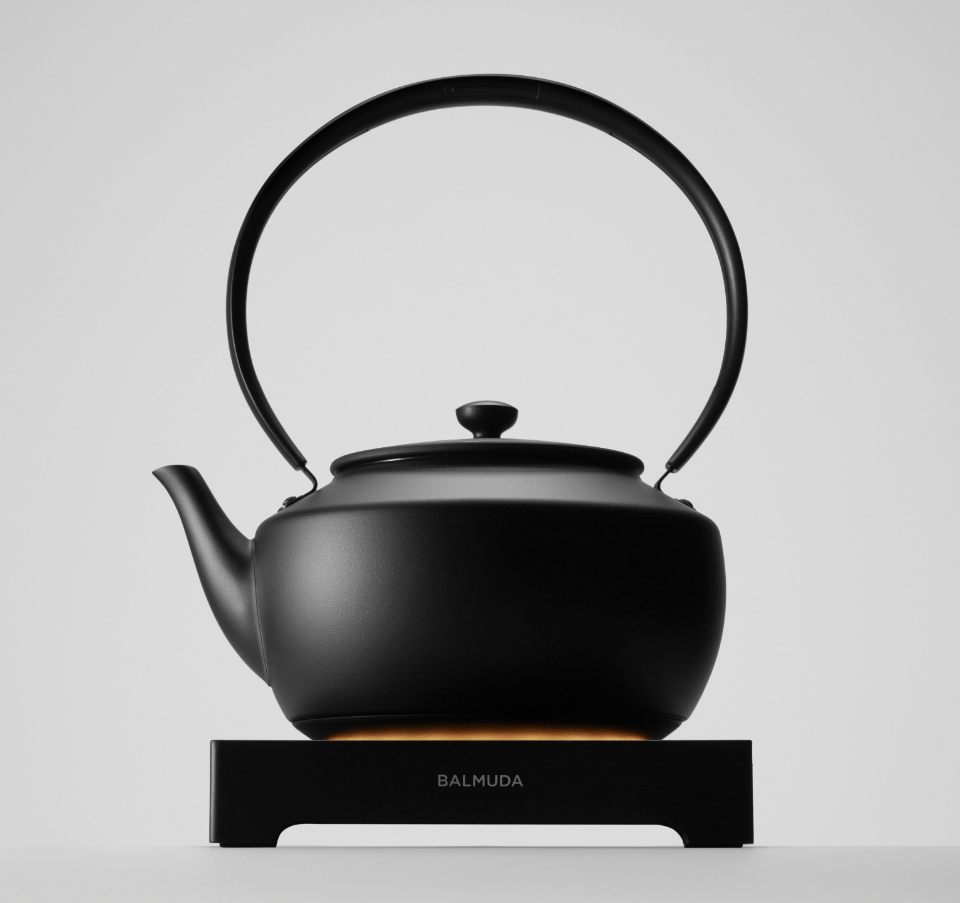 A matte black BALMUDA MoonKettle resting on its base, glowing subtly beneath, against a minimalist white background.