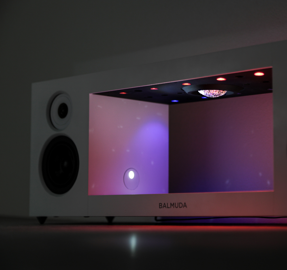 Close-up of an early prototype of a BALMUDA speaker with a built-in projection or lighting feature, showing glowing lights and reflections inside the inner compartment. Like a live stage with lights about and speakers on the outside of the stage.