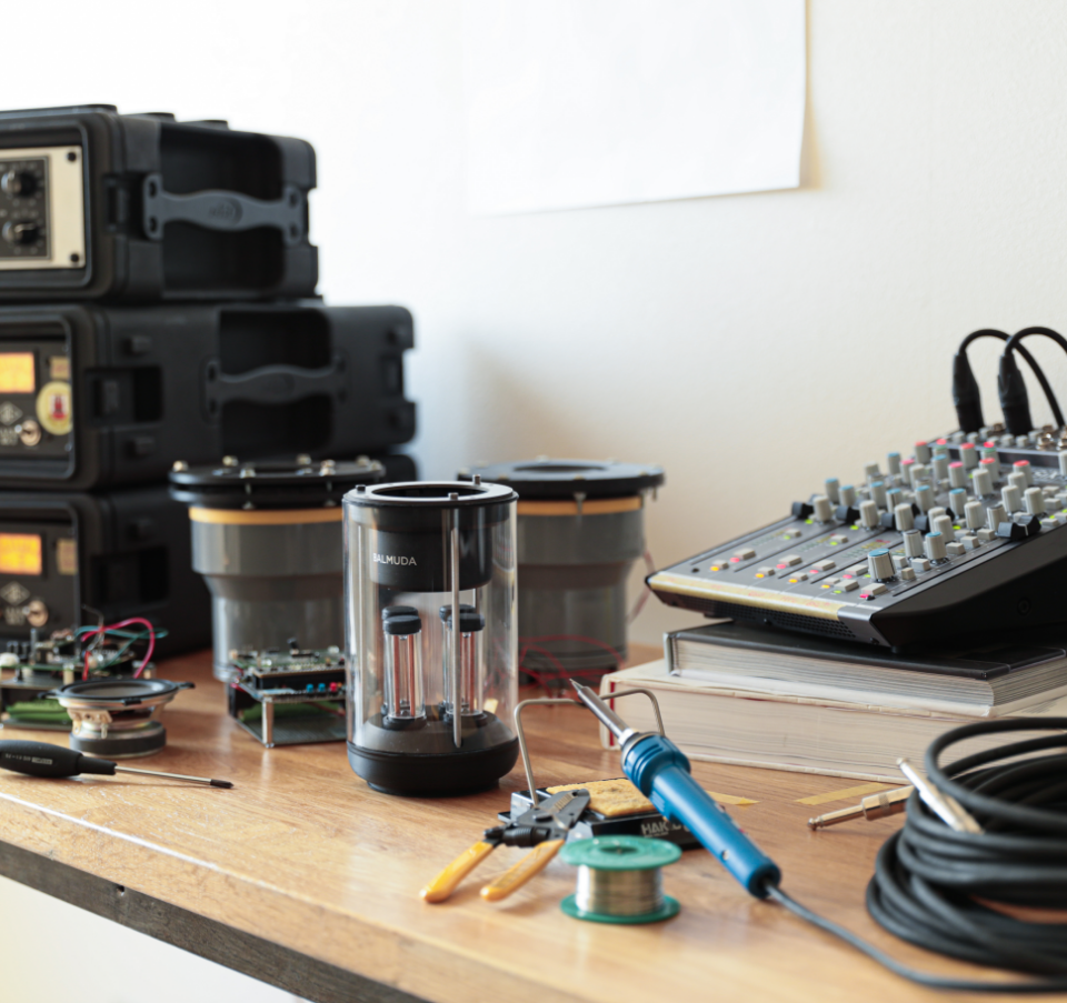 A speaker prototype placed on a wooden workbench surrounded by audio components, soldering tools, and a sound mixer.