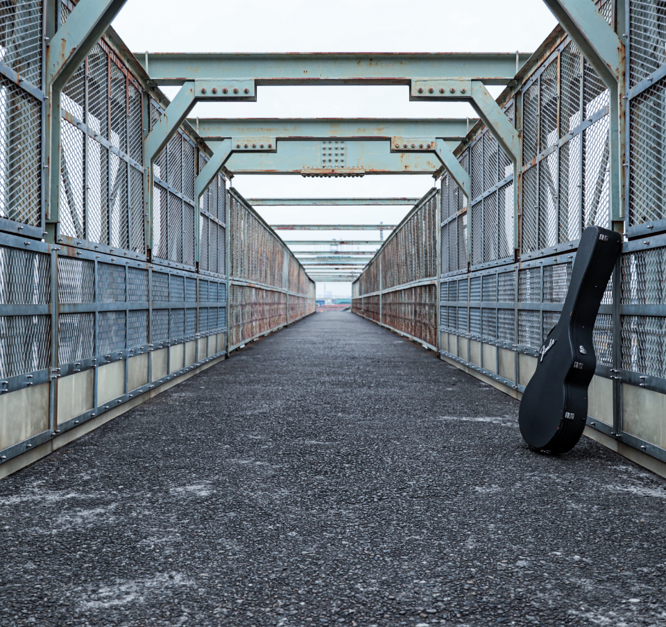 A black guitar case standing upright against a metal fence on an empty pedestrian bridge with industrial-style steel beams and a textured asphalt walkway.