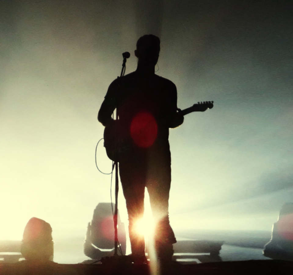 Silhouette of a musician playing electric guitar on stage with a microphone stand, backlit by intense stage lights and surrounded by fog or haze.