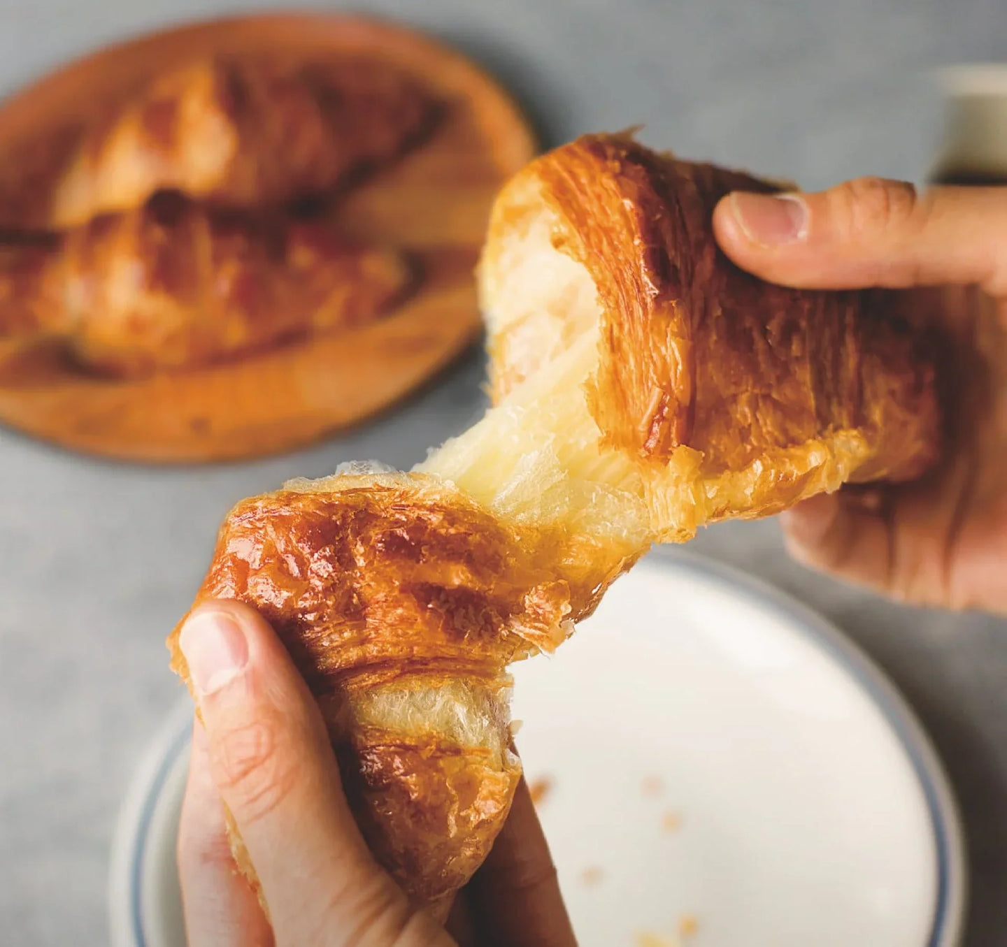 Golden-brown croissant being split in half, showing its crispy exterior and soft inside, with more croissants on a wooden plate in the background. That was warmed in The Toaster