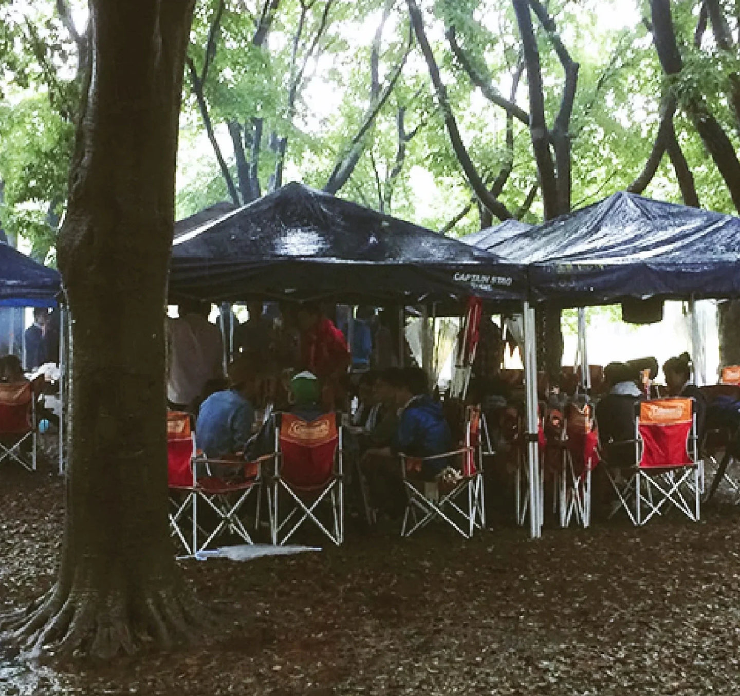 group of people camping in the rain under black tents in a wooded area. They are seated in red and orange folding chairs, chatting and staying dry.
