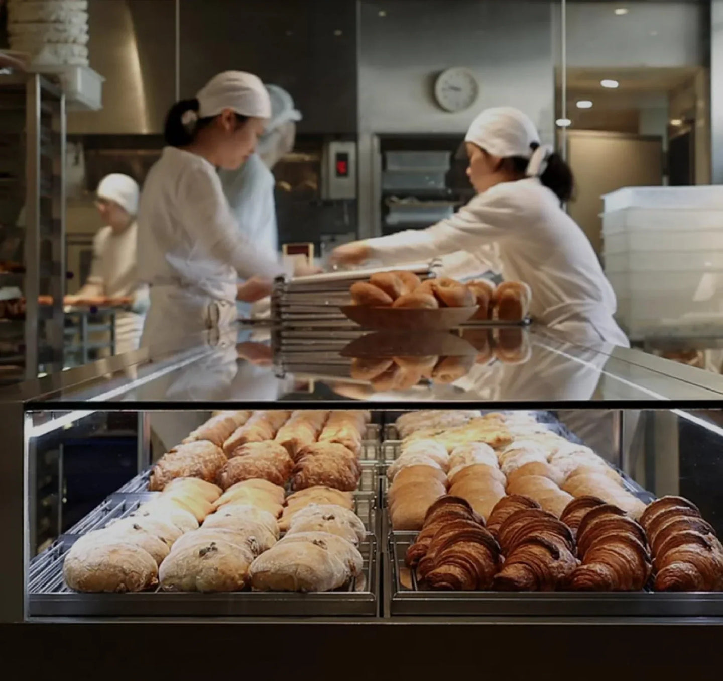 A bakery kitchen with workers dressed in white uniforms and head coverings, preparing fresh pastries. The foreground showcases a glass display case filled with golden croissants and a variety of bread, while behind the counter, bakers arrange and handle baked goods in a modern kitchen setting.
